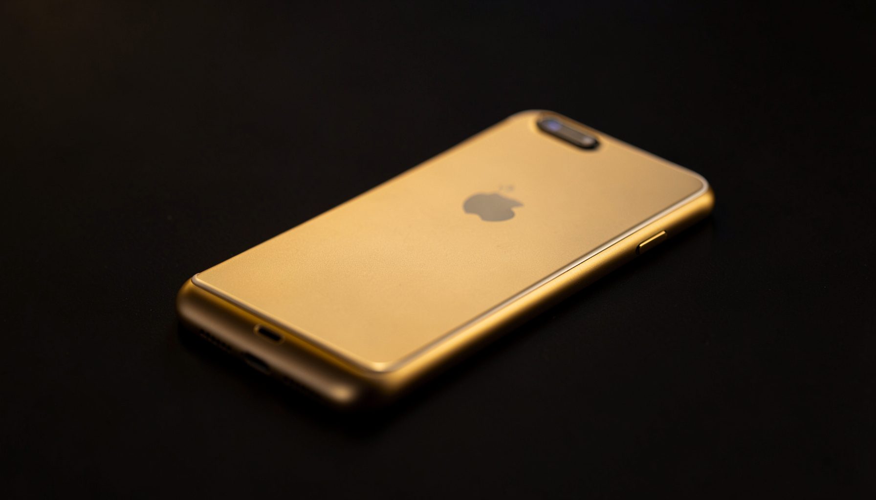 Smartphone dengan bodi ramping berwarna emas keemasan di atas permukaan gelap, cahaya studio hangat menyorot dari samping.